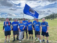 A group of adults in blue baseball jerseys stand on a field with a flag behind them that reads Claws Out
